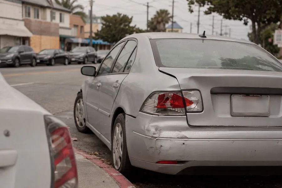 Rear end collision damage and a broken taillight on a silver sedan parked at the curb